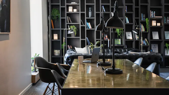 Et poleret bord har sorte lamper og en bærbar computer. Omkring det står sorte stole. I baggrunden, en lædersofa og en bogreol med bøger og planter skaber en hyggelig biblioteksstemning.