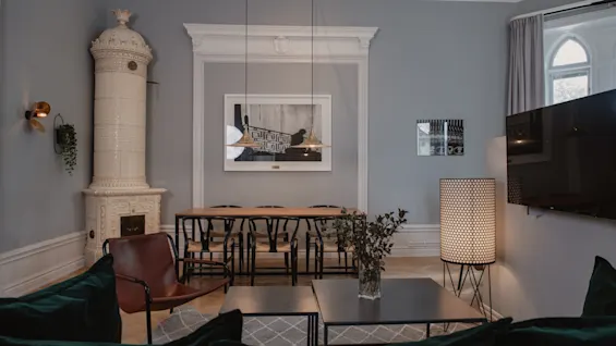 Ett vardagsrum har en modern, mysig inredning med en rund vit keramisk kamin, en brun läderstol och ett matbord med stolar. Rummet har en grå vägg, en stor målning, en golvlampa och en TV.
