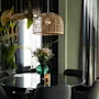 Et stilig spiseområde med et rundt svart bord, fire stoler, en stor plante, en taklampe i rotting, blomster og et lys.