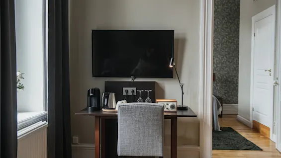 Et skrivebord med en grå stol har en kaffemaskine, kedel og lampe under et vægmonteret TV. Beliggende i et rum med vindue og et delvist synligt tilstødende område.