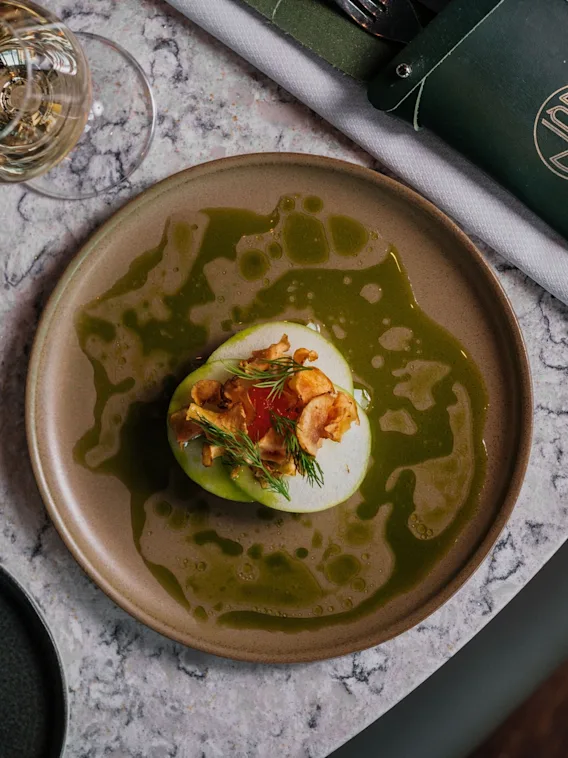 En elegant anrettet rett med grønn saus, hvit base, sprø topping og friske urter, sammen med vinglass.