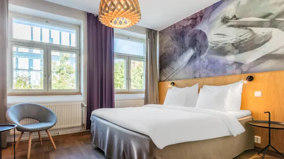 A modern bedroom with a neatly made bed, wooden headboard, and a large mural above. The room features a window, cozy chair, and pendant lamp. "BAGARE" is written on the mural.