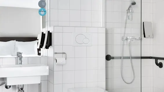 Ett modernt badrum med vita kakel, ett handfat med spegel, en dusch med handmunstycke och en toalett med dubbelspolningsknappar. Skyltar lyder "GO EASY ON THE PLANET" och "DRINK MORE WATER."