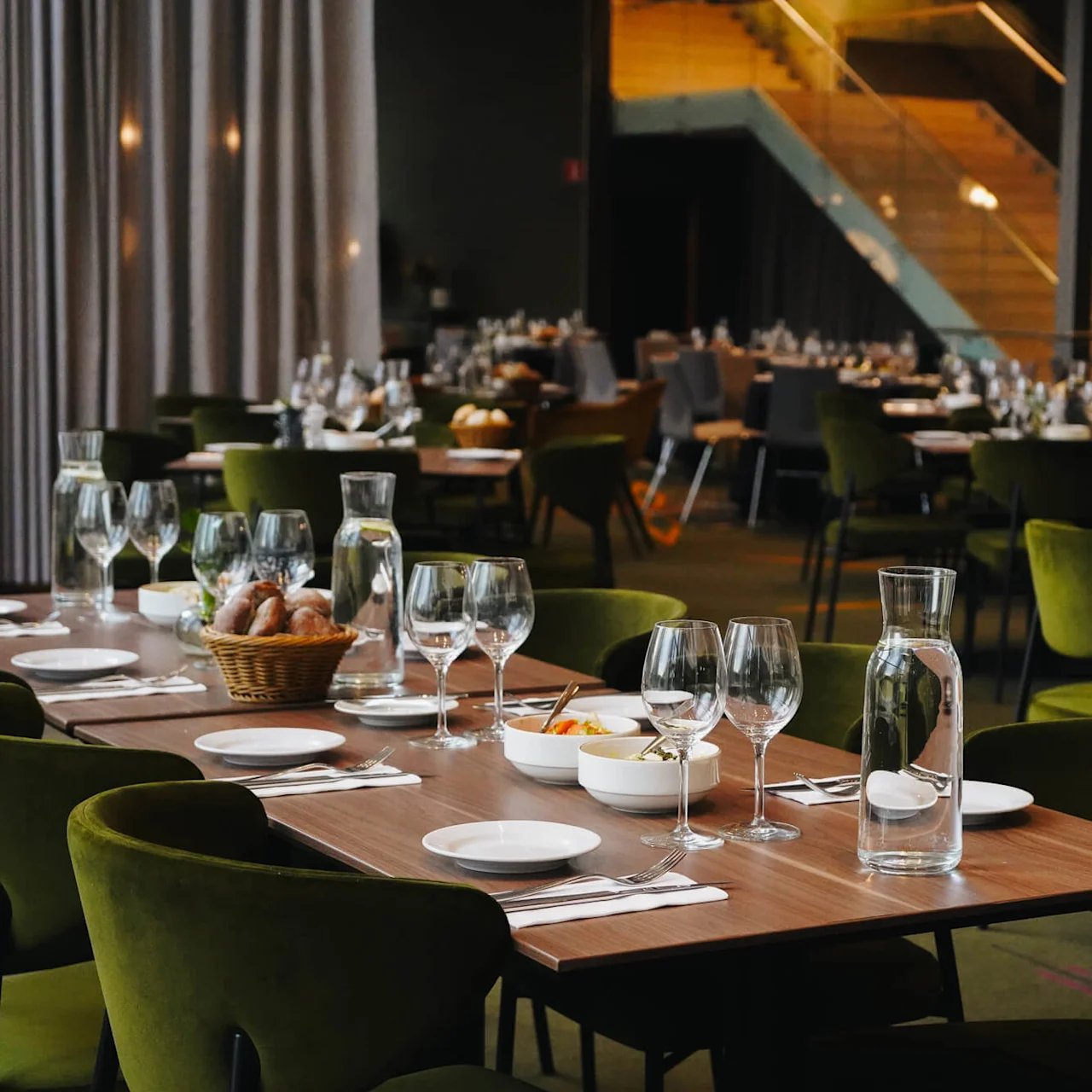 Elegant dining tables set with glassware, bowls, and napkins. Green chairs surround the tables in a softly lit restaurant. Staircase and draped curtains in the background.