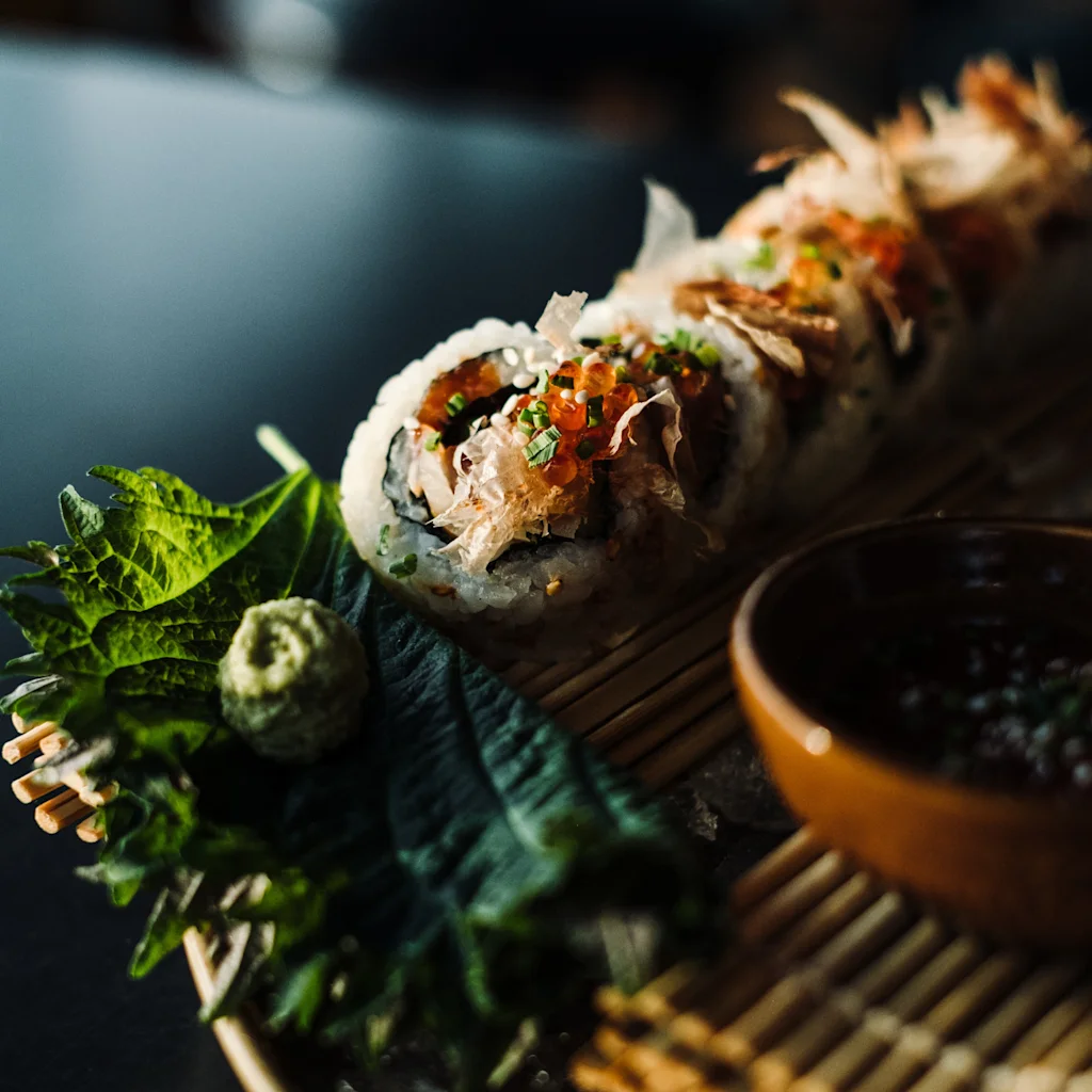 En smukt præsenteret sushirulle, garneret med levende toppings, ligger på en bambusmåtte sammen med friske grøntsager og en dyppesauce.