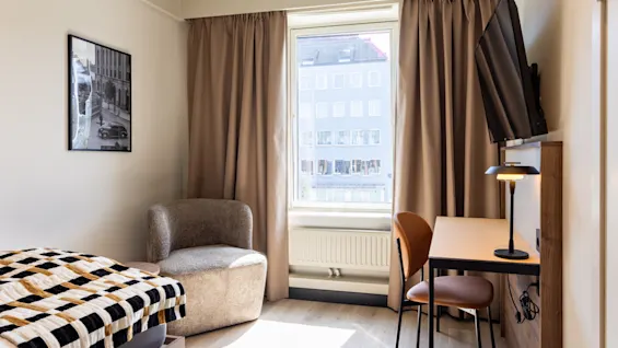 Et koselig hotellrom med seng, en komfortabel stol, et skrivebord og et vindu med utsikt over byen.