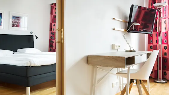 A desk with a chair stands near a wall, facing a mounted TV. Nearby, a bed with white linens is partially visible. Red curtains decorate the scene. 