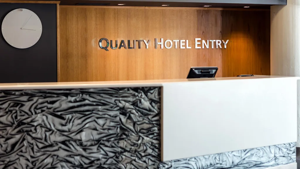En hotellresepsjon med moderne design står under en trevegg med teksten "QUALITY HOTEL ENTRY." En hvit klokke henger på en mørk bakgrunn.