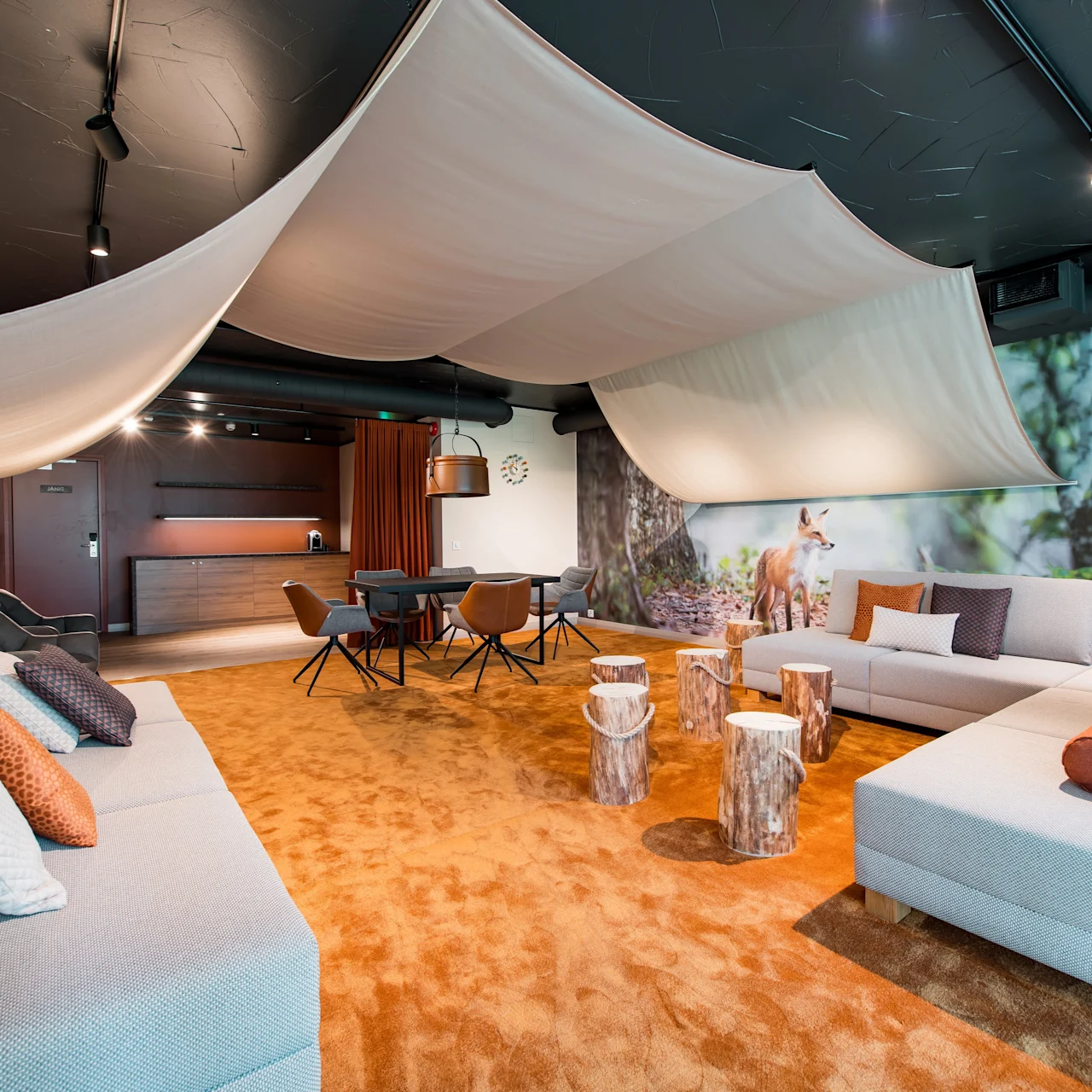 Luxurious lounge with sofas, draped fabric ceiling, and wooden stump tables in a room featuring forest-themed wall art. Warm lighting and earthy tones create a cozy atmosphere.