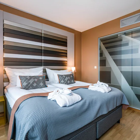 En luksuriøs seng er pænt arrangeret med puder og foldede morgenkåber, mod et dekorativt panel i et moderne hotelværelse, ved siden af en gennemsigtig glasvæg, der fører til et flisebelagt badeværelse.