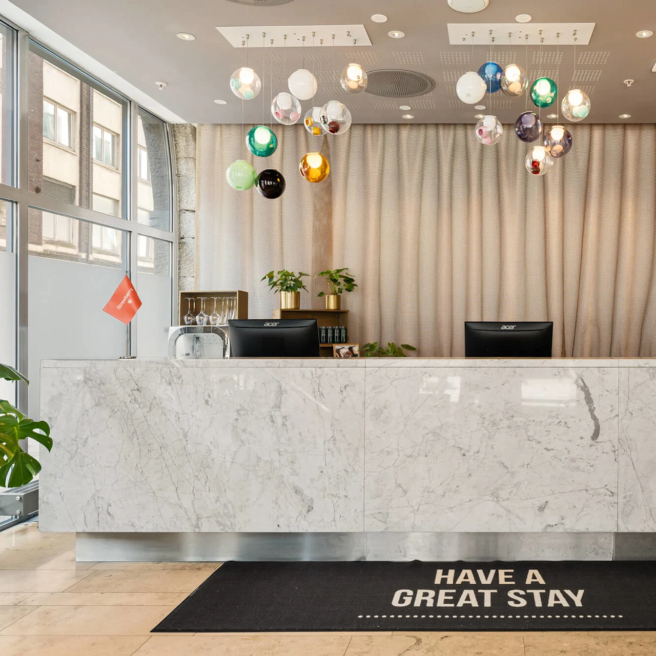 Receptionen står med computere, planter og dekorative lys ovenfor. En sort måtte på gulvet lyder "HAVE A GREAT STAY.”