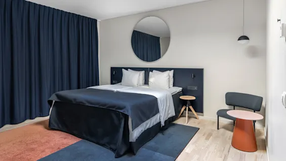 Et moderne hotelværelse med en stor seng, mørkeblå gardiner, et rundt spejl og moderne møbler.