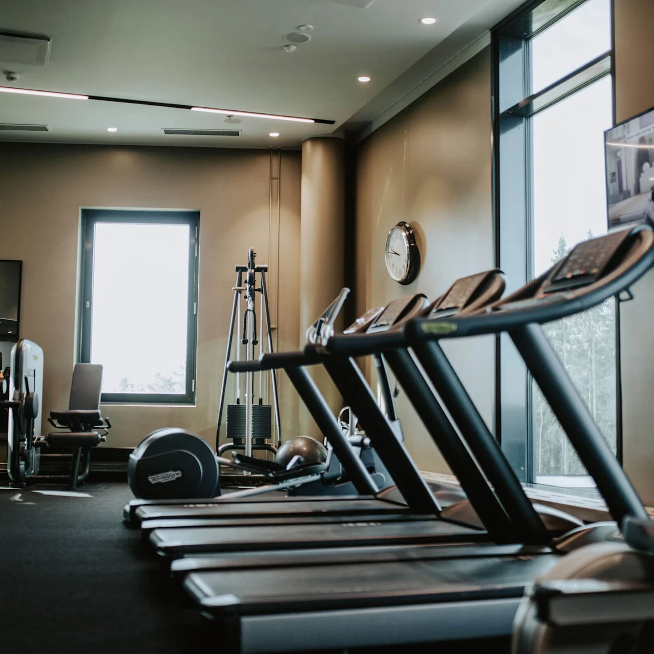 Treadmills are positioned in a brightly lit gym with mirrors, exercise machines, a wall clock, and a window showing an outdoor view. A television displays people exercising.