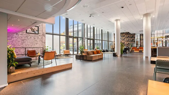 A spacious, modern lobby features stylish seating arrangements and large floor-to-ceiling windows providing natural light. Plants and contemporary decor enhance the aesthetic, creating a welcoming atmosphere.