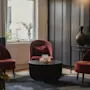 Et hyggeligt hotelloungeområde med tre røde lænestole, et sofabord med stearinlys og et vindue med naturligt lys.