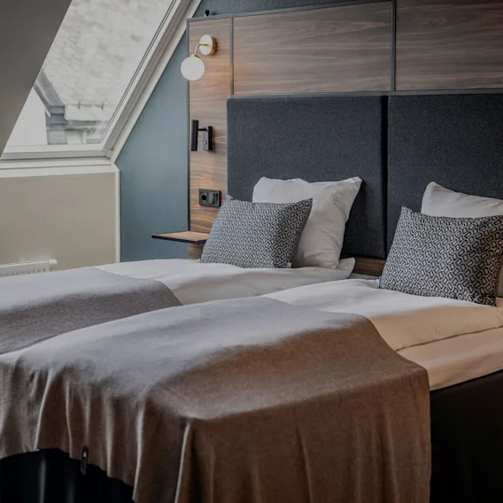 Hotellrom med to enkeltsenger redd opp i grått og hvitt sengetøy under et skråstilt takvindu.