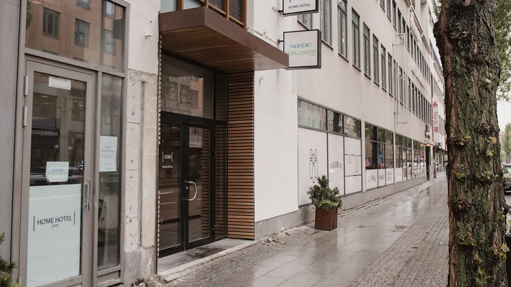 A hotel entrance with glass doors labeled "Home Hotel" and "Odin's Hotel & Spa" adjacent to a city sidewalk, flanked by a tree and a lined row of windows.