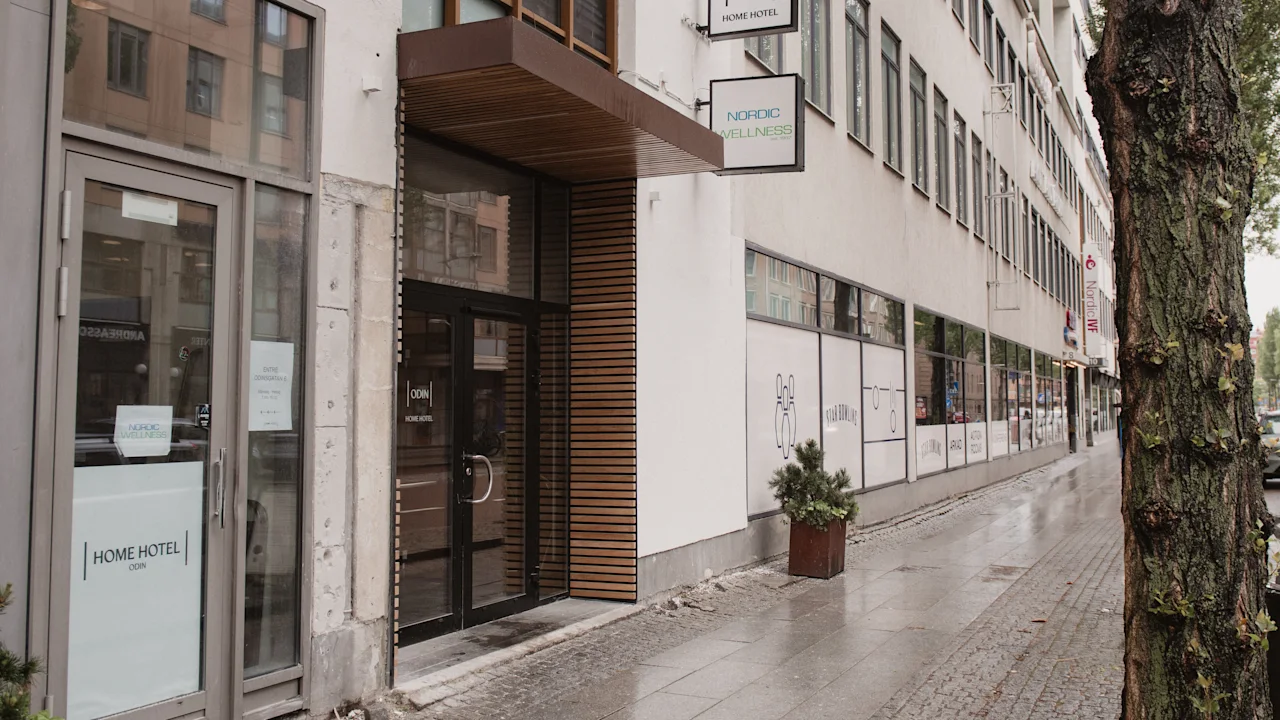 Ingång med glasdörrar märkta "Home Hotel" och "Odin's Hotel & Spa" vid en stadstrottoar, omgiven av ett träd och en rad fönster.