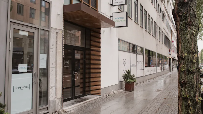 A hotel entrance with glass doors labeled "Home Hotel" and "Odin's Hotel & Spa" adjacent to a city sidewalk, flanked by a tree and a lined row of windows.