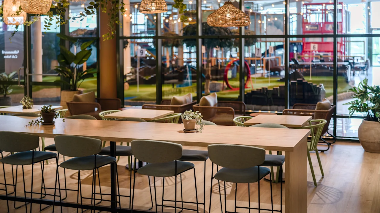 Et langt trebord med krakker står i en moderne innendørskafé, dekorert med hengende lamper, planter og store vinduer som avslører et gresskledd uteområde.