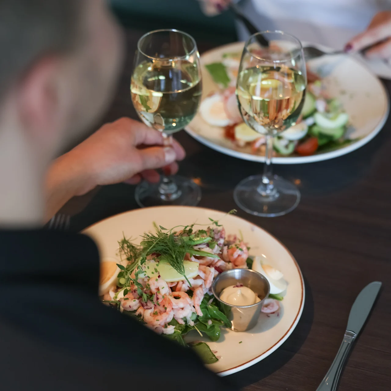 A seafood salad garnished with herbs is served on a white plate, accompanied by a glass of white wine, on a wooden dining table set for two people.