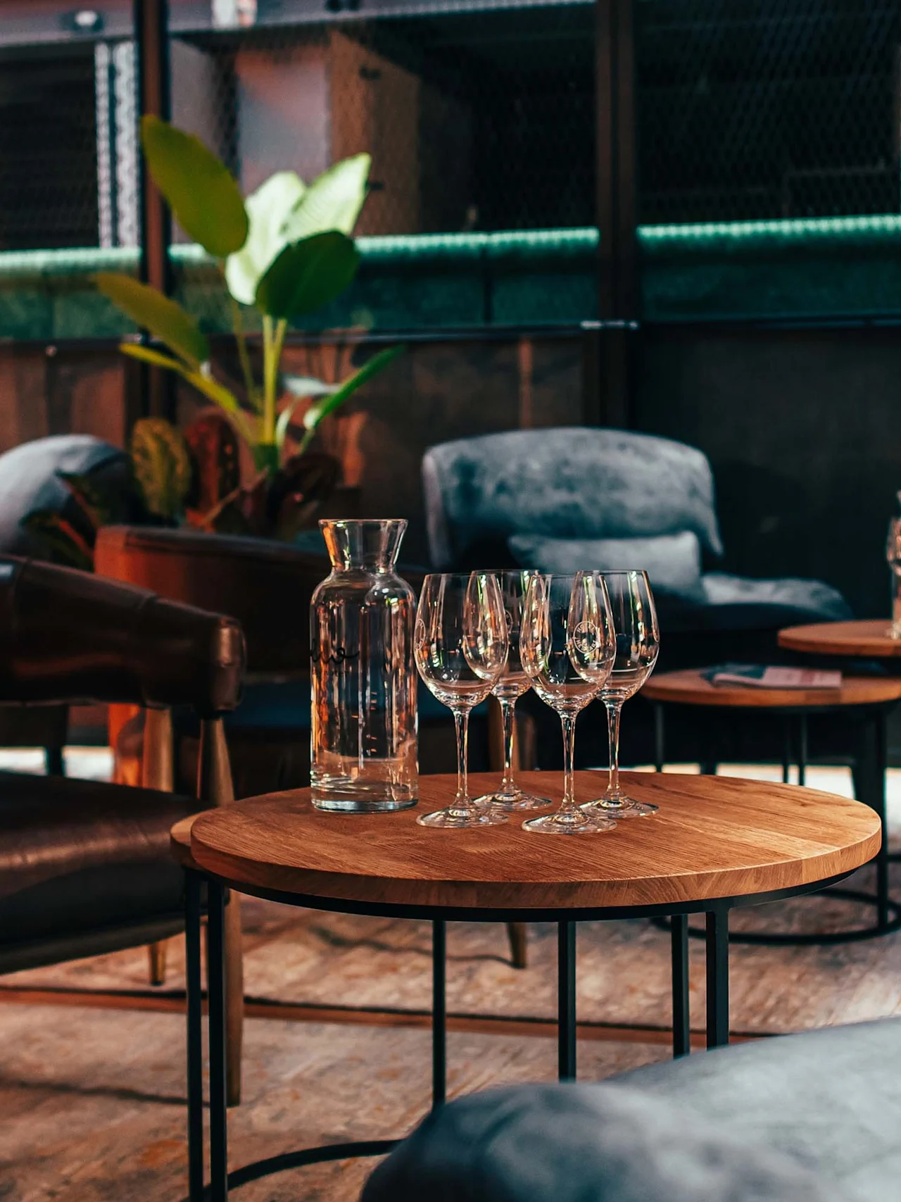 Water carafe and wine glasses rest on wooden coffee tables surrounded by leather armchairs in a cozy, warmly lit lounge. Lush potted plants and soft upholstered chairs provide a relaxing ambiance.