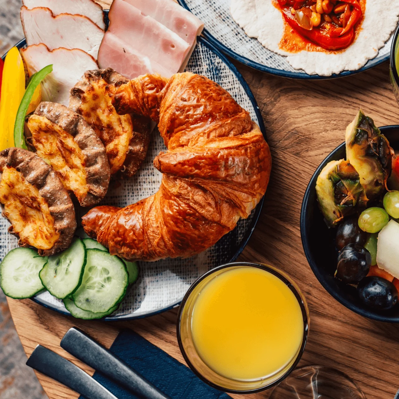 Croissant, skinke og osteboller ligger på et bord med skivede grønnsaker, fruktsalat, appelsinjuice, grønn smoothie og kaffe rundt.