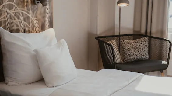 Viihtyisä hotellihuone, jossa sänky valkoisilla liinavaatteilla, vehnäkuvioitu seinä, tyylikäs nojatuoli ja lattiavalaisin.