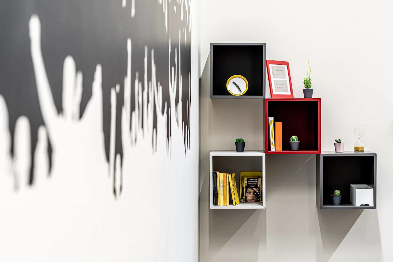 Wall-mounted cube shelves display books, small plants, a clock, and a framed picture. A black-and-white mural of silhouettes decorates the adjacent wall.