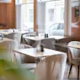 Bord og stoler er pent plassert i en koselig kafé med dempet belysning. Store vinduer slipper inn naturlig lys, med utsikt over parkerte biler utenfor.