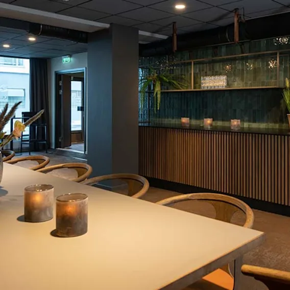 Et langt bord med lys og blomster står i en moderne kafé, omgitt av trestoler. En disk med planter og en kaffemaskin pryder veggen.