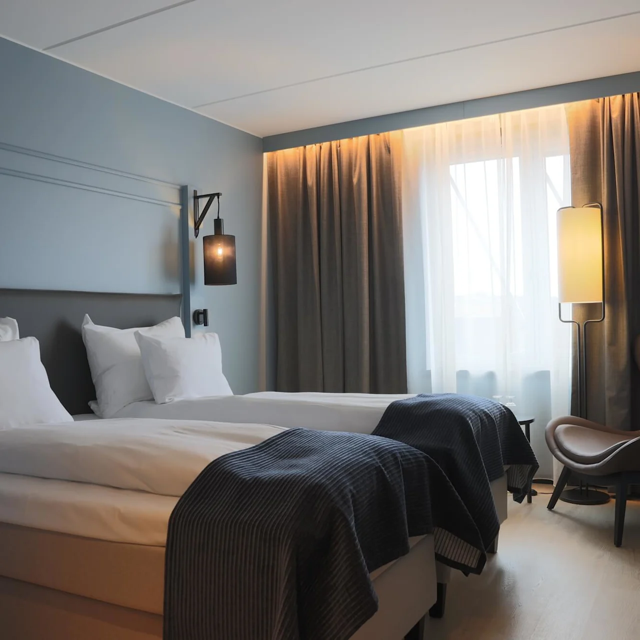 A neatly made bed in a modern hotel room with soft lighting, curtains drawn, and a cozy chair by the window.