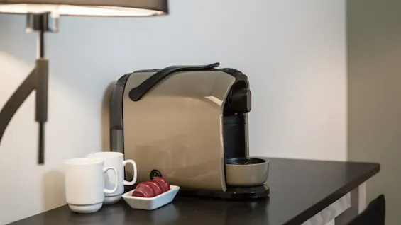 A coffee machine sits on a black countertop, accompanied by two white mugs and a dish holding three red coffee pods. A lamp is partially visible above.