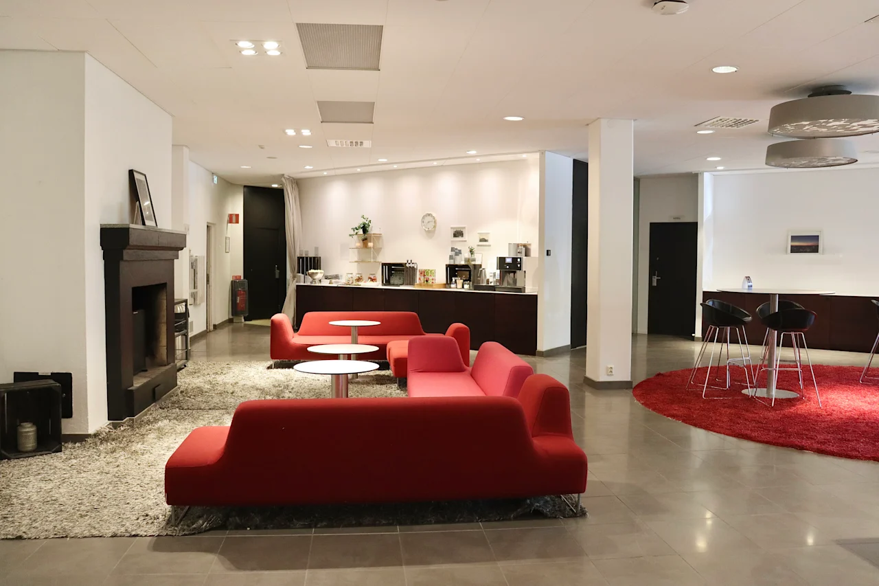 Red sofas encircle round tables in a spacious, modern lobby. A fireplace and coffee station add comfort, while high stools and tables sit on a circular red rug nearby.