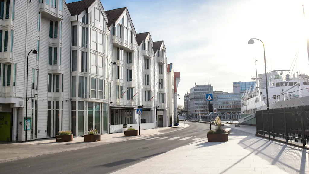 En solrig byscene med elegante hvide bygninger langs en gade, med en havn og et stort fartøj i det fjerne.