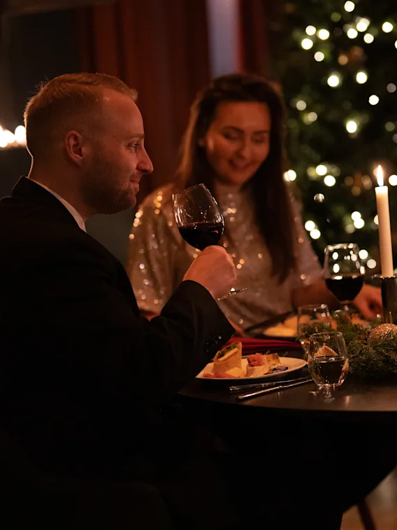 Gäster njuter av en festlig middag med vin och levande ljus, med en dekorerad julgran i bakgrunden.