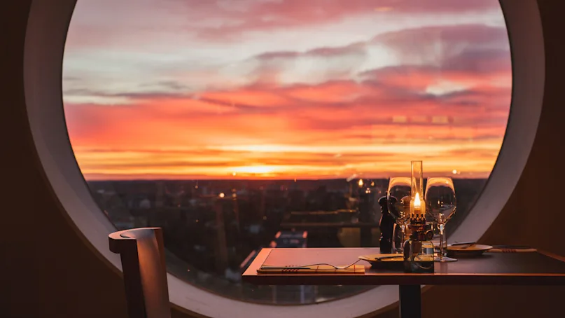 A table set for dinner with a candle and glasses is positioned near a large round window, with a vibrant sunset casting hues of orange and pink in the sky.