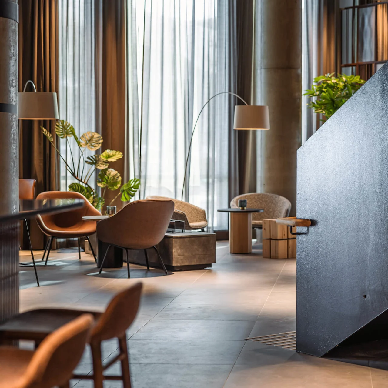 Et moderne loungeområde har stilfulde stole arrangeret omkring lave borde, med en elegant sort trappe til højre og store vinduer med tynde gardiner i baggrunden.