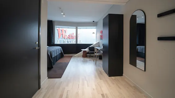 A hotel room features a bed, desk, and mirror, with a window displaying a "Westfield Mall of Scandinavia" sign.