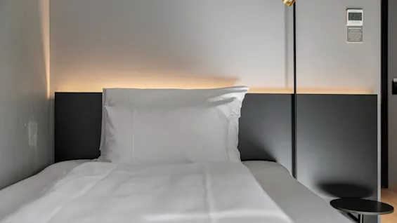 En pent oppredd seng med hvitt sengetøy og pute, opplyst av et varmt skjær bak sengegavlen, innbyr til et avslappende opphold.