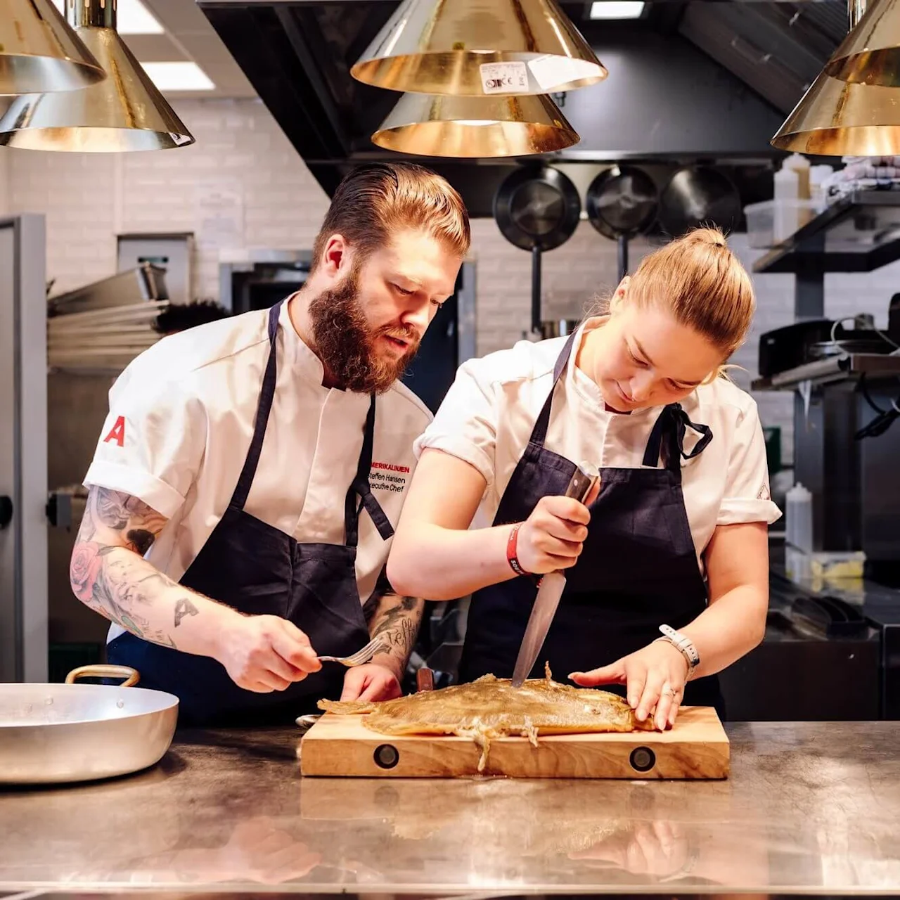 Two chefs in a commercial kitchen prepare a fish on a wooden board under warm lighting. One uses a large knife, while the other assists with a fork.