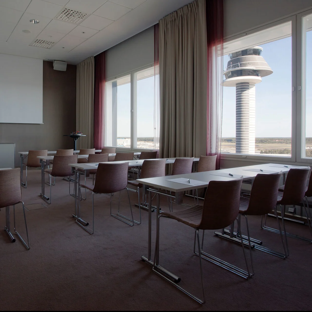 A classroom features rows of empty chairs and tables facing a large white projection screen; outside the window, an airport control tower is visible, surrounded by a clear sky.
