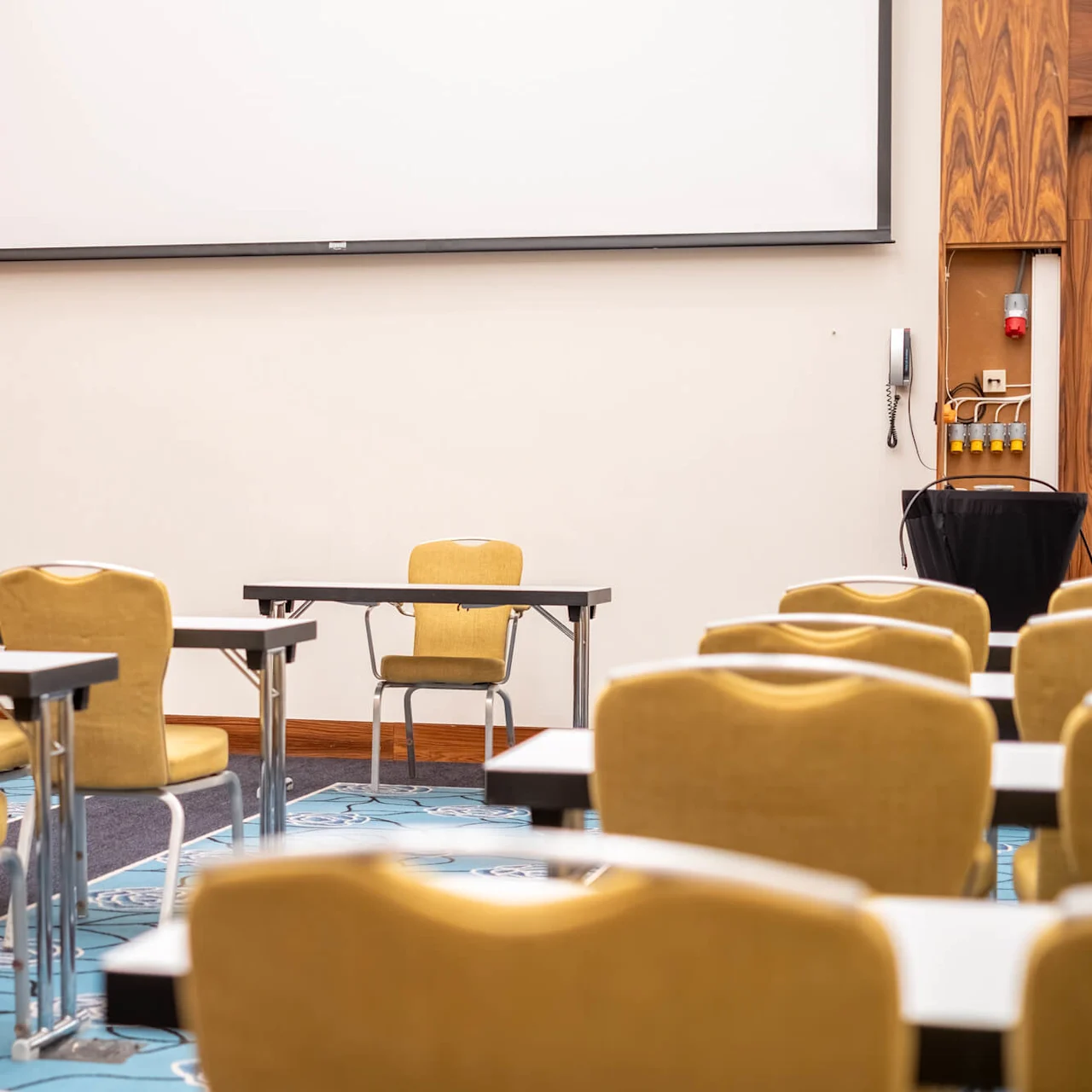 Empty conference room features rows of yellow chairs facing a large blank projection screen. Wooden paneling and a patterned carpet complete the setting.