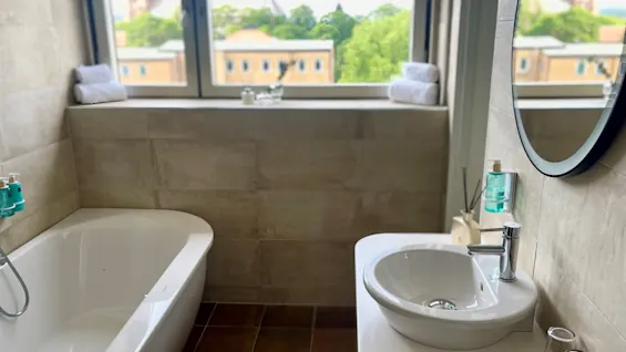 A white bathtub and sink sit in a modern bathroom. Two towels rest on a windowsill overlooking a cityscape with trees and buildings in the background.