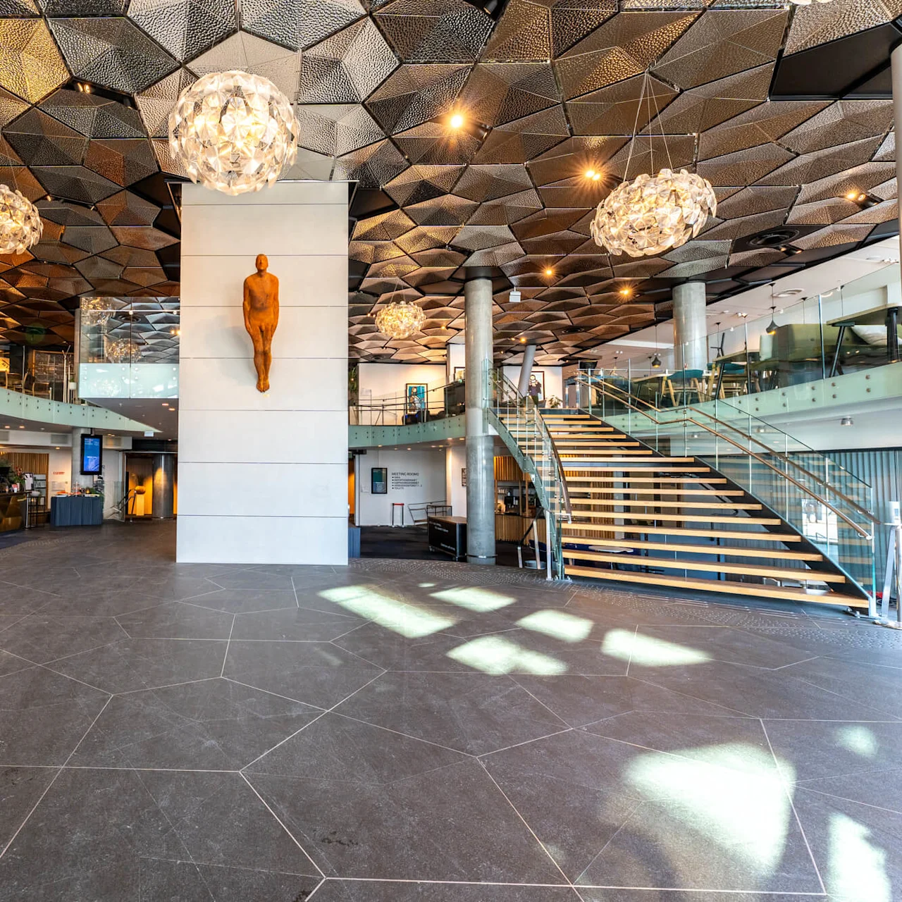 Lobby med moderne loftdetaljer og lysekroner. Receptionen er til venstre, trappe til højre. En skulptur er monteret på væggen. Tekst: "CLARION HOTEL THE EDGE."