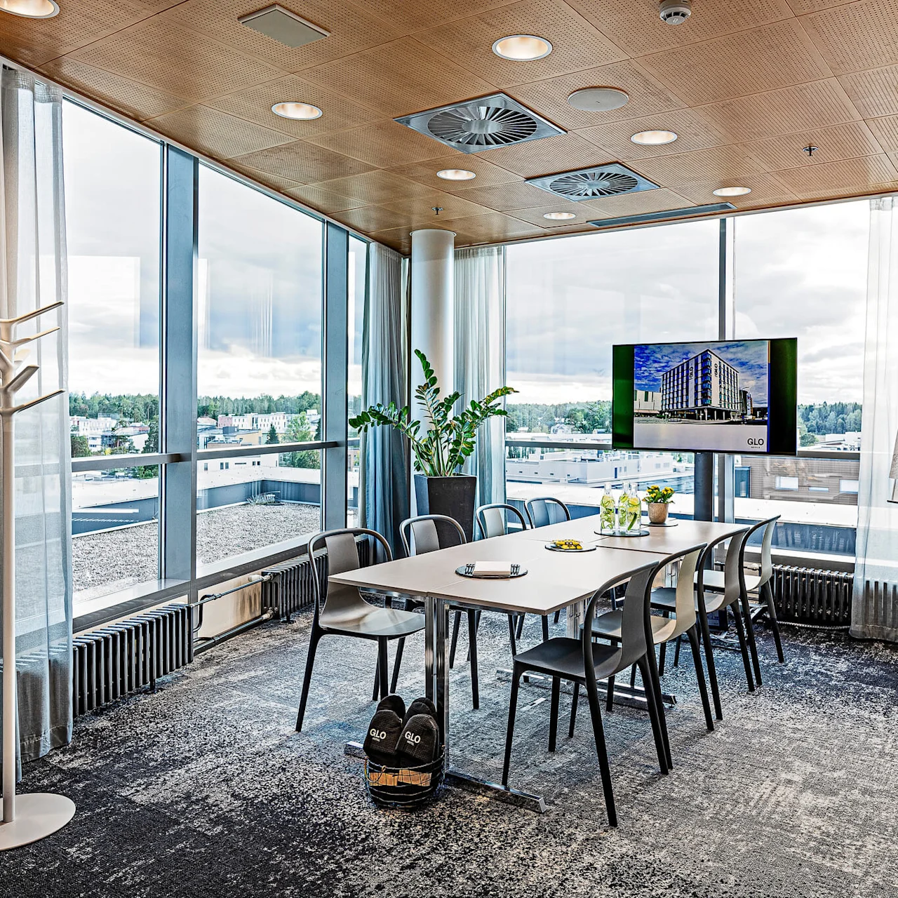 Conference table surrounded by chairs, situated in a glass-walled room with urban views. A presentation board reads, "GLO, GREAT TO HAVE YOU HERE." A TV shows a building image.