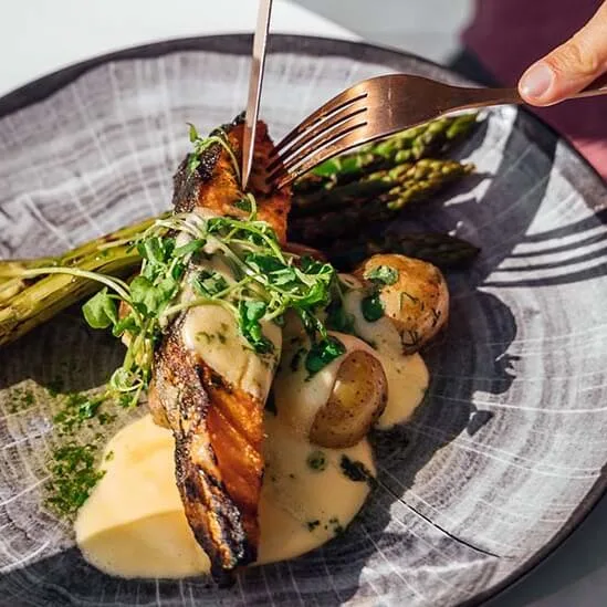 En tallerken med grillet laks, asparges, poteter og saus; en person skjærer i fisken med en gaffel.