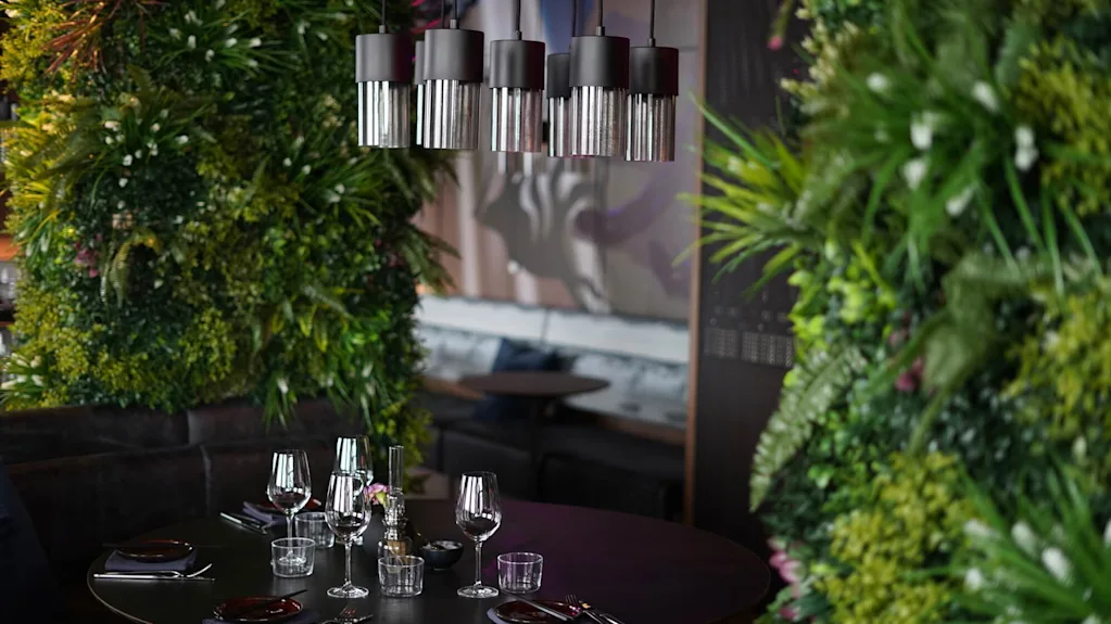 Moderna hänglampor hänger över ett runt matbord med glas och bestick, omgivna av frodiga gröna väggar i en mysig, exklusiv restaurangmiljö.