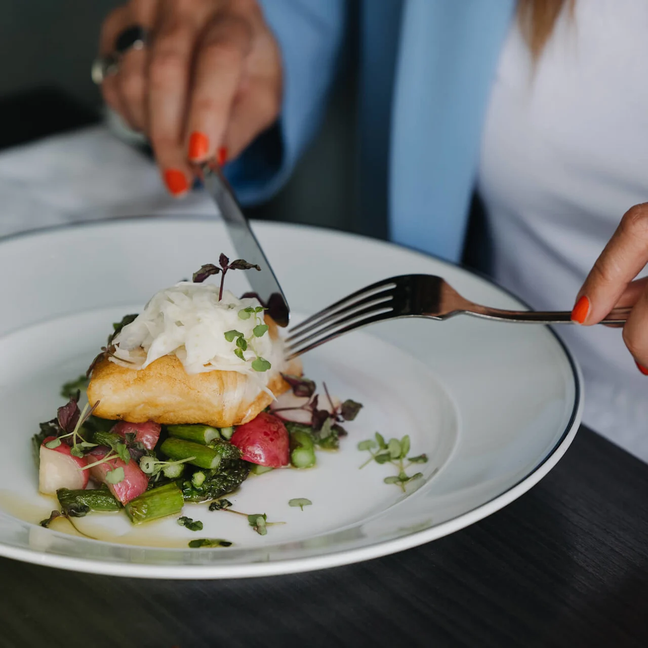 En tallerken med kokt fisk toppet med grønt serveres med grønnsaker. En person med røde negler skjærer den med bestikk ved siden av et glass hvitvin på et bord.