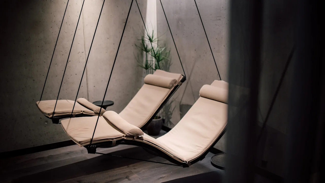 Two beige lounge chairs, suspended from the ceiling, hang in a minimalist room with dark wooden floors and concrete walls. A small side table and a potted plant are nearby.